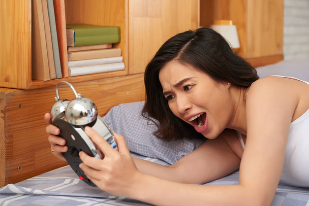 Sobredormida joven asiática mirando su despertador con la boca abierta
  - Foto, imagen