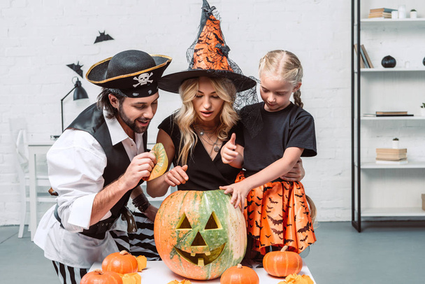Porträt von Eltern und kleiner Tochter in Halloween-Kostümen, die zu Hause gemeinsam in den Kürbis schauen - Foto, Bild