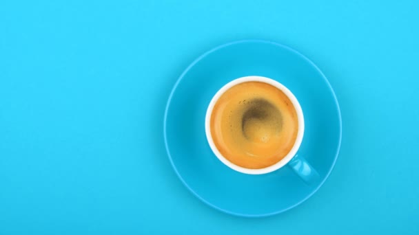 Close up one full white cup of espresso coffee and saucer over pastel blue paper background with slow motion animated spin of coffee froth, elevated top view, directly above - Footage, Video