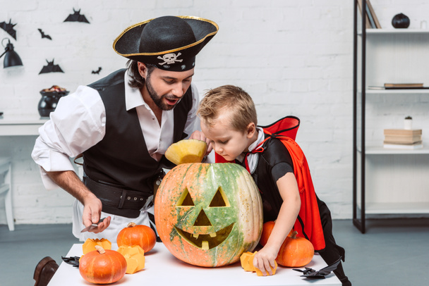man in pirate costume and son in vampire halloween costume looking into pumpkin together at home - Foto, Imagem