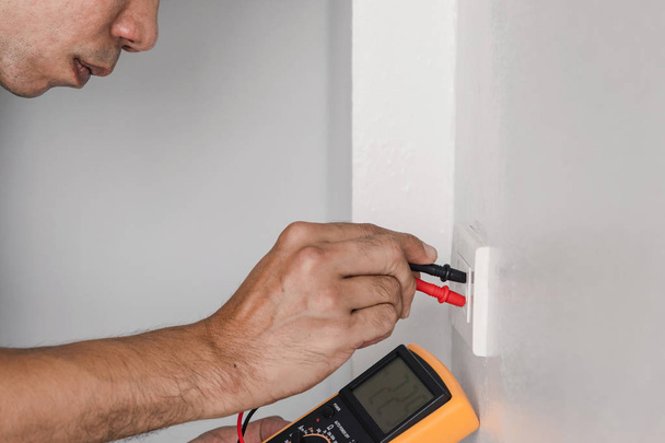 Электрик использует цифровой счетчик для измерения напряжения в розетке питания на стене
. - Фото, изображение