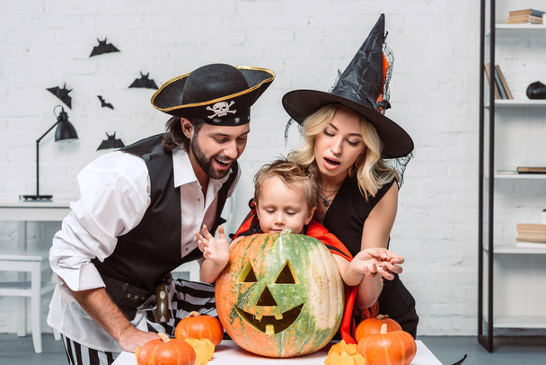 portrait of parents and son in halloween costumes looking into pumpkin together at home - Photo, Image
