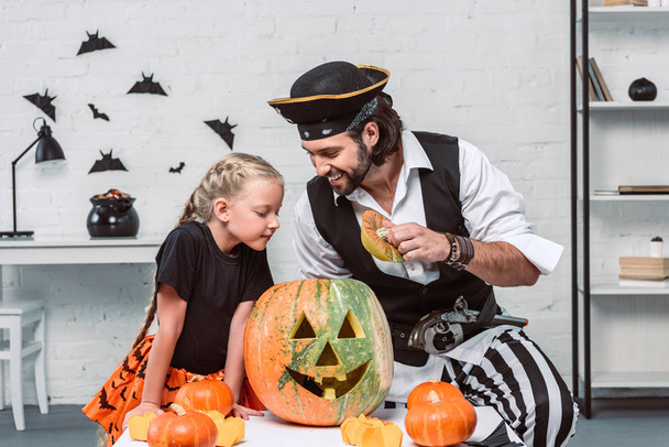 father and daughter in halloween costumes looking into pumpkin together at home - Foto, Bild
