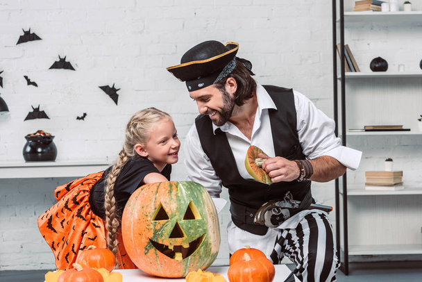father and daughter in halloween costumes looking into pumpkin together at home - Φωτογραφία, εικόνα
