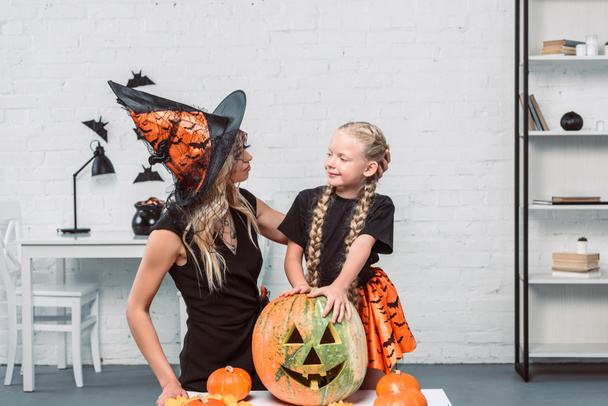portrait of mother and little daughter in witches costumes at coffee table with pumpkins at home - Fotoğraf, Görsel