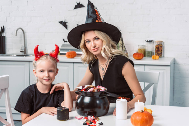 мати і маленька дочка в костюмах Хеллоуїна за столом з частуваннями в чорному горщику на кухні
 - Фото, зображення