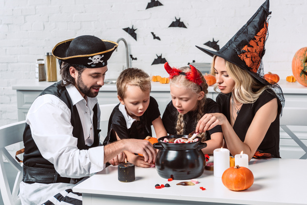 familie in verschillende kostuums van halloween aan tafel met zwarte pot met behandelt in keuken thuis - Foto, afbeelding