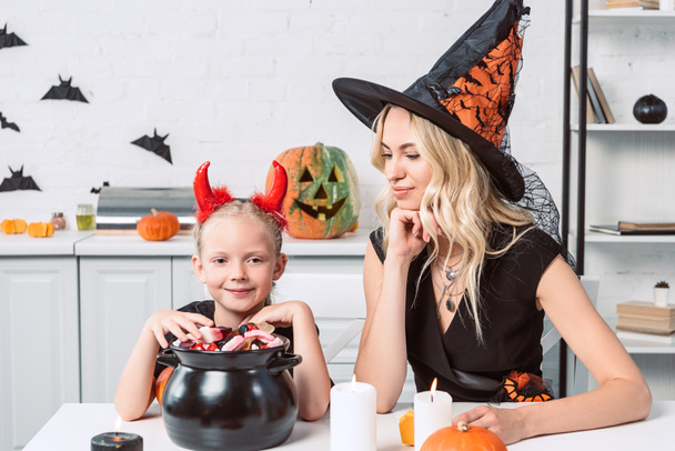 Mutter und kleine Tochter in Halloween-Kostümen am Tisch mit Leckereien im schwarzen Topf in der Küche - Foto, Bild