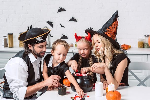 Familie in verschiedenen Halloween-Kostümen am Tisch mit schwarzem Topf mit Leckereien in der heimischen Küche - Foto, Bild