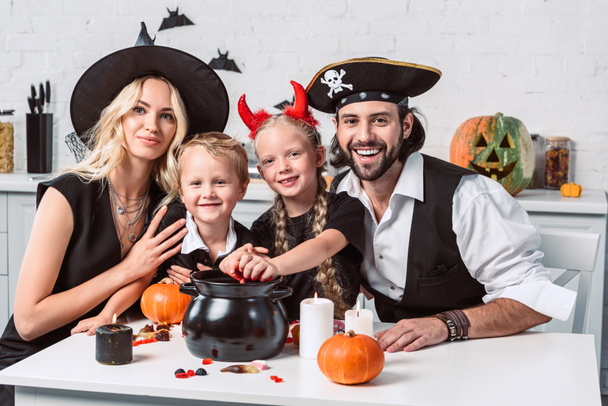 family in various halloween costumes at table with black pot with treats in kitchen at home - Photo, Image