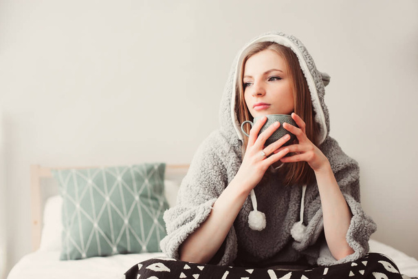 young beautiful woman relaxing at home on the bed, wearing funny grey hoodie. Chilling in lazy weekend morning with cup of tea or coffee - Foto, Imagem