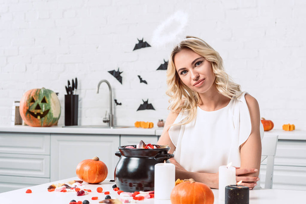 portrét krásné ženy v kostýmu anděl sedící u stolu s sladkosti v černý hrnec pro halloween - Fotografie, Obrázek