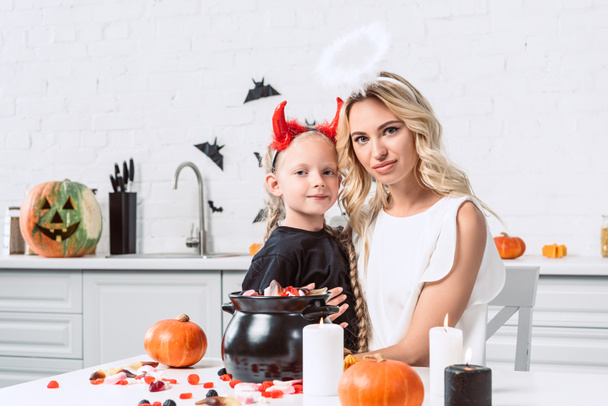 Portret van de moeder en dochter in de kostuums van halloween aan tafel met zoetigheden in zwarte pot in de keuken thuis - Foto, afbeelding