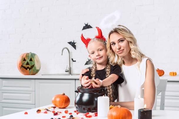portrait of mother and daughter in halloween costumes at table with sweets in black pot in kitchen at home - Photo, Image