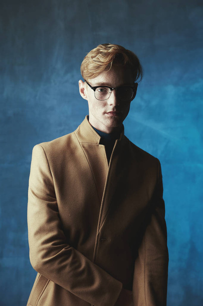 Dark studio portrait of young male model with red hair and freckles. Weared in brown coat and glasses. Blue light and background. - Foto, imagen