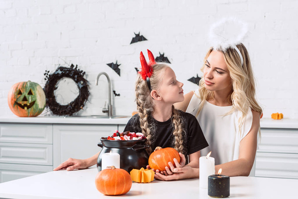 portrait of mother and daughter in halloween costumes at table with sweets in black pot in kitchen at home - Foto, Imagem