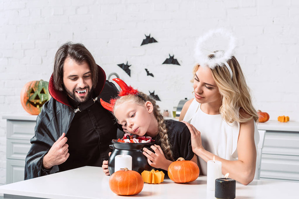 portrait of parents and daughter in halloween costumes at table with treats in black pot in kitchen at home - Foto, afbeelding
