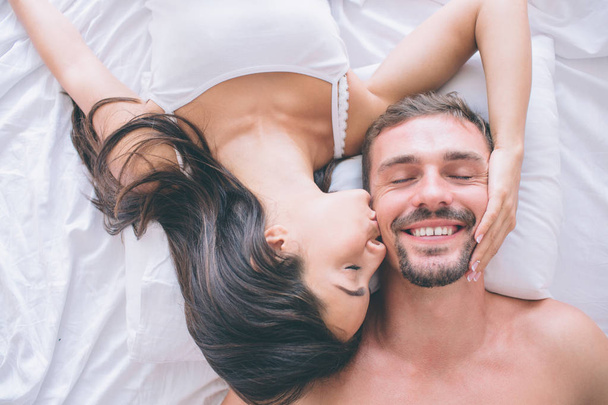Gorgeous and attractive couple is lying in bed face to face. Guy keeps his eyes closed and smiles. Girl holds her hand on his cheek and kisses him. - 写真・画像