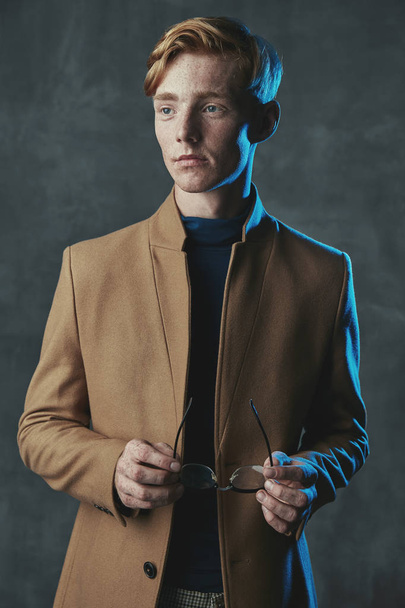 Studio portrait of young male model with red hair and freckles. Weared in brown coat, with glasses in hand. Blue light, gray background. - Foto, Imagem