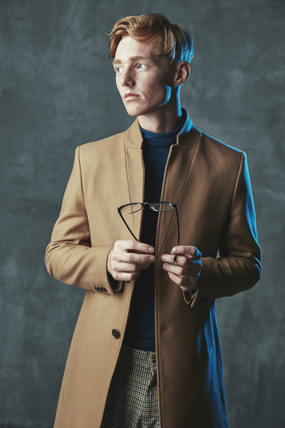 Studio portrait of young male model with red hair and freckles. Weared in brown coat, plaid pants, with glasses in hand. Blue light, gray background. - Фото, зображення