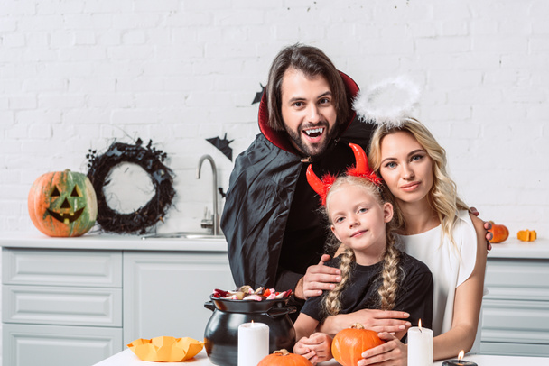 portrait of parents and daughter in halloween costumes at table with treats in black pot in kitchen at home - Фото, зображення