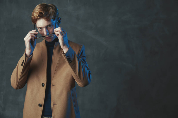 Studio portrait of young male model with red hair and freckles. Dressed in brown coat, wearing glasses. Blue light, gray background. - Фото, изображение