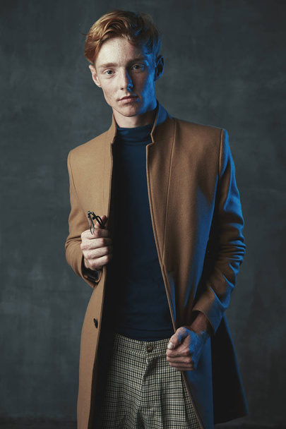 Studio portrait of young male model with red hair and freckles. Weared in brown coat, plaid pants, with glasses in hand.  - Foto, imagen