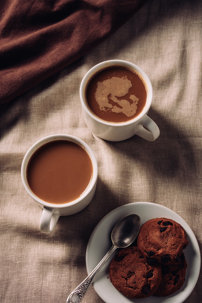 top view of cups of coffee with chocolate chip cookies on beige cloth - 写真・画像