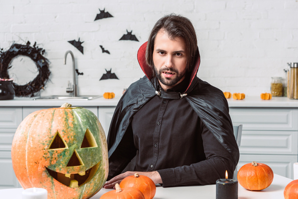 Portret van man in vampier halloween kostuum zittend aan tafel met pompoenen in keuken thuis - Foto, afbeelding