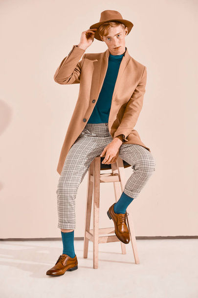 Young male model with red hair and freckles posing in brown coat and hat, plaid pants, turquoise sweater and socks, brown shoes, sitting on chair. Pink background  - Фото, изображение