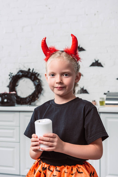 portrait of little kid with red devil horns holding candle in hands at home, halloween holiday concept - Fotó, kép