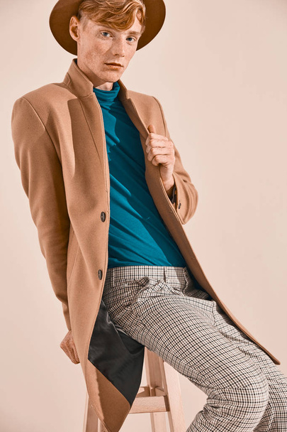 Young male model with red hair and freckles in brown coat and hat, plaid pants, turquoise sweater, sitting on chair. Creamy background  - Foto, imagen