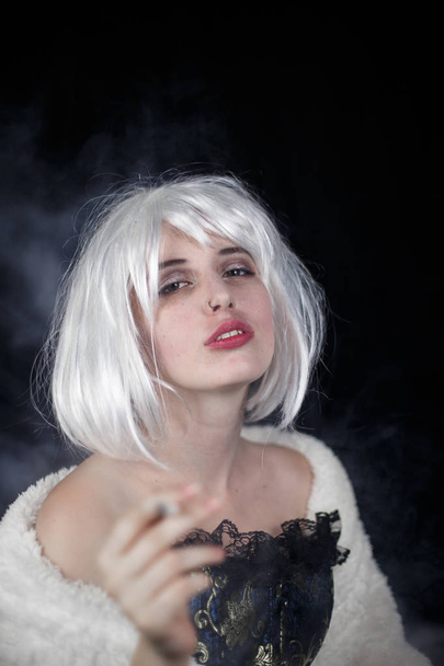 Atractiva joven mujer en traje de fumar cigarro y mirando a la cámara mientras está de pie sobre fondo negro
 - Foto, Imagen