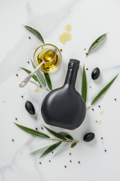 κάτοψη του μαύρο μπουκάλι λάδι ελιάς και ποτήρι με κουτάλι στο μαρμάρινο τραπέζι - Φωτογραφία, εικόνα