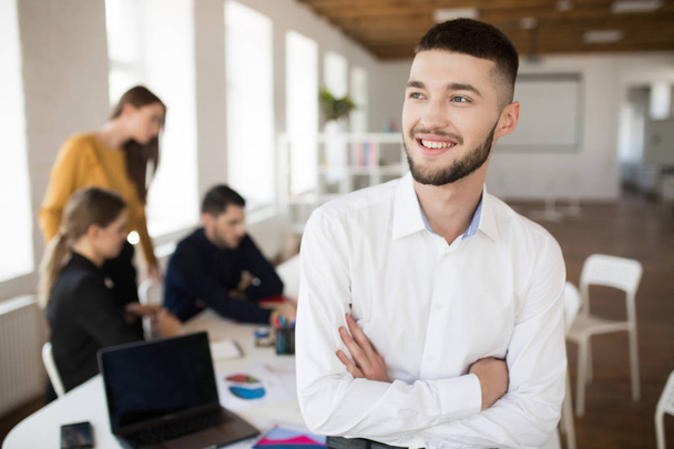 Молодий усміхнений чоловік з бородою в білій сорочці щасливо виглядає осторонь зі складеними руками, проводячи час в офісі з колегами на фоні
 - Фото, зображення