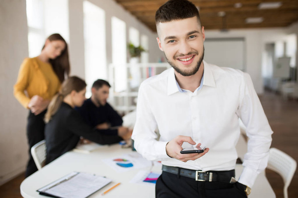 Молодой улыбающийся мужчина с бородой в белой рубашке счастливо смотрит в камеру, держа мобильник в руке, проводя время в офисе с коллегами на заднем плане
 - Фото, изображение