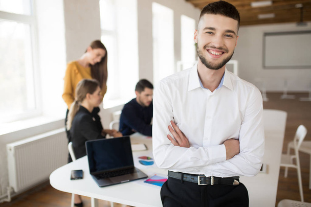 Молодой улыбающийся мужчина с бородой в белой рубашке счастливо смотрит в камеру со сложенными руками, проводя время в офисе с коллегами на заднем плане
 - Фото, изображение