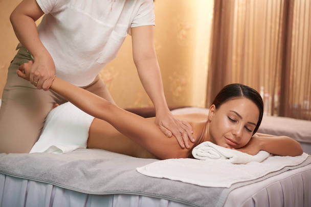 Young beautician stretching arm of relaxed female during body massage in spa salon - Photo, Image