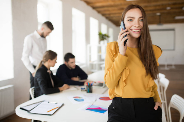 Pretty smiling woman happily looking in camera talking on cellphone in office with colleagues on background - Photo, Image