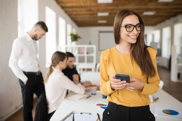 Молодая деловая женщина в очках радостно смотрит в сторону, держа мобильный телефон в руках в офисе с коллегами на заднем плане
 - Фото, изображение