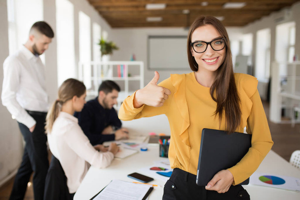 Fiatal mosolygó üzleti nő boldogan keresi a kamera találat hüvelykujjával felfelé gesztus mappát a Hivatal munkatársaival kezében háttér szemüvegek - Fotó, kép
