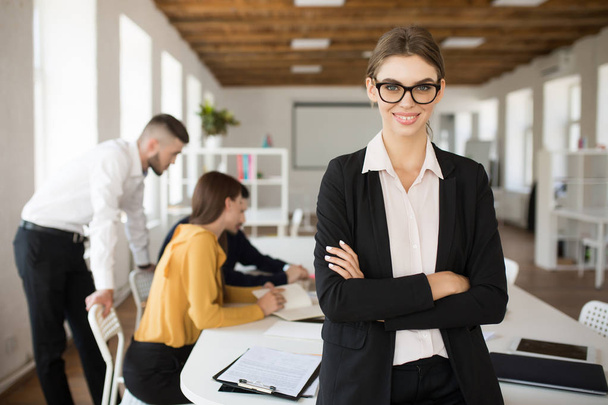 Young smiling business woman in eyeglasses and shirt happily looking in camera with folded hands spending time in office with colleagues on background - Foto, Imagen
