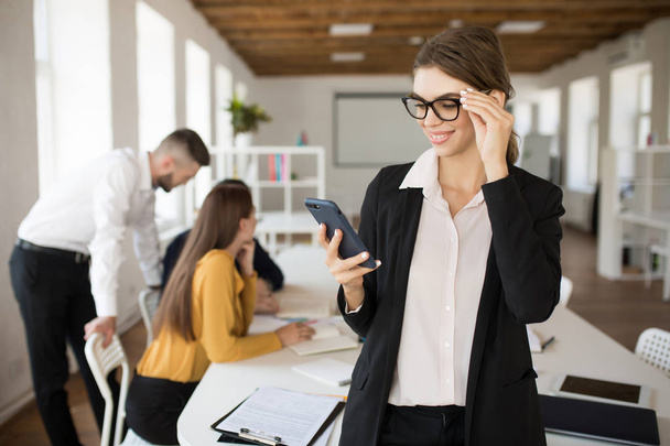 Молодая улыбающаяся деловая женщина в очках и рубашке с удовольствием использует мобильный телефон проводить время в офисе с коллегами на заднем плане
 - Фото, изображение
