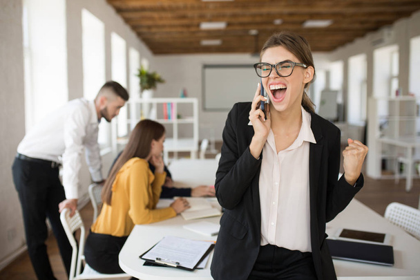 Jonge zakenvrouw in brillen en shirt emotioneel schreeuwen op mobiel in kantoor in gesprek met collega's op achtergrond - Foto, afbeelding
