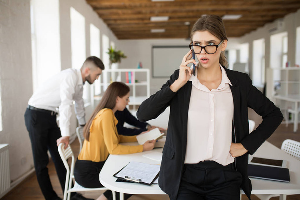 Young serious business woman in eyeglasses and shirt amazedly looking aside talking on cellphone in office with colleagues on background - Foto, Imagen