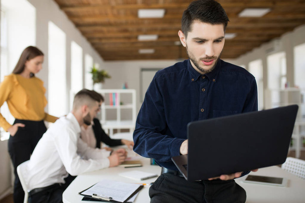 Fiatal férfi szakáll póló elgondolkodva használ laptop tölteni az időt a Hivatal munkatársaival a háttérben - Fotó, kép