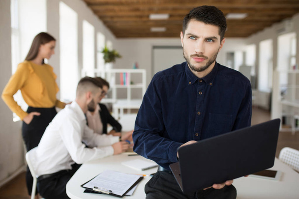 Jovem homem sério com barba na camisa olhando atenciosamente de lado segurando laptop em mãos passar tempo no escritório com colegas no fundo
 - Foto, Imagem