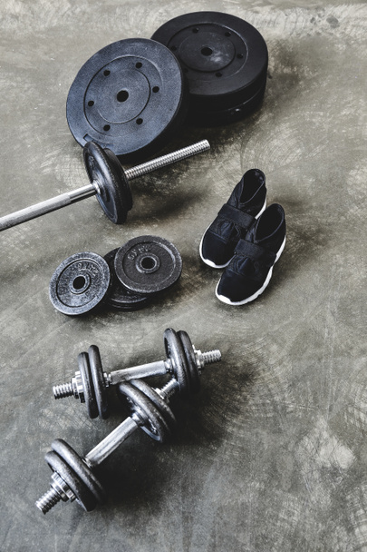 dumbbells and barbell with weight plates and sneakers on grungy grey floor - Fotó, kép