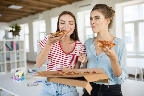 Two pretty girls sitting on desk joyfully eating big pizza spending time together in modern office - Foto, Bild