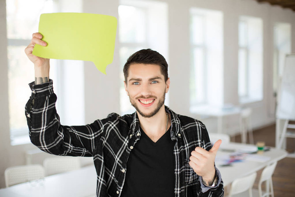 Young smiling man with beard happily looking in camera showing thumb up holding green paper in cloud shape in hand near head in modern office - Fotoğraf, Görsel